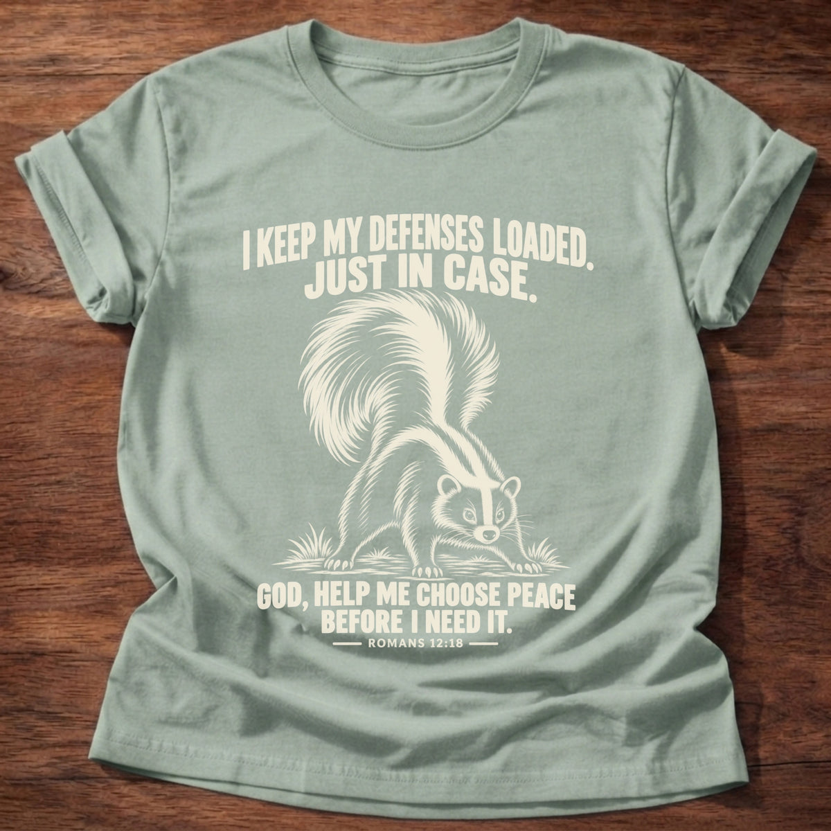 Sage t-shirt printed with a skunk with its tail raised, ready to spray graphic and 'I keep my defenses loaded. Just in case. God, help me choose peace before I need it.' Romans 12:18 faith based gift