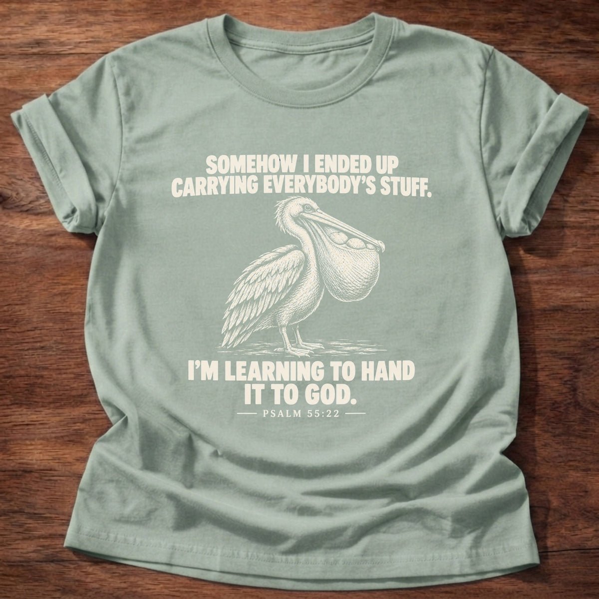 Sage t-shirt printed with a pelican with a pouch full of fish graphic and 'Somehow I ended up carrying everybody's stuff. I'm learning to hand it to God.' Psalm 55:22 faith based gift