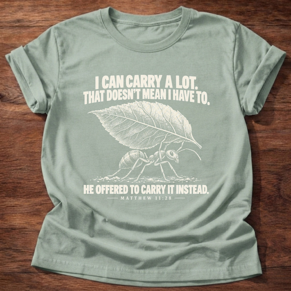 Sage t-shirt printed with an ant hauling a leaf many times its size graphic and 'I can carry a lot. That doesn't mean I have to. He offered to carry it instead.' Matthew 11:28 faith based gift