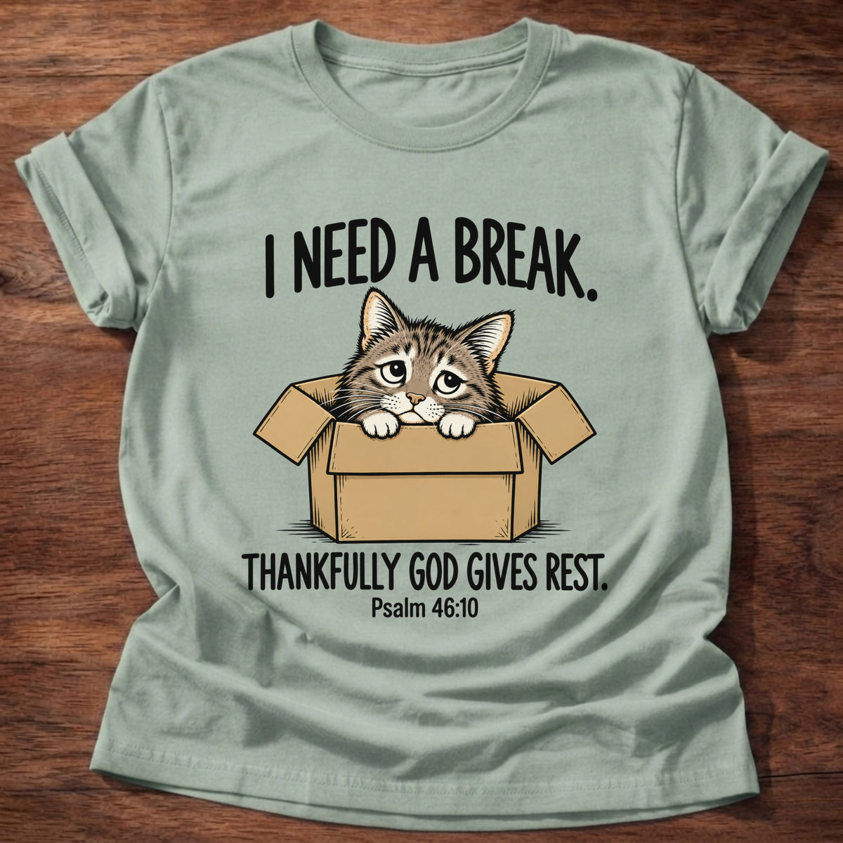 Sage t-shirt printed with a cat curled in a cardboard box graphic and 'I need a break. Thankfully God gives rest.' Psalm 46:10 faith based gift
