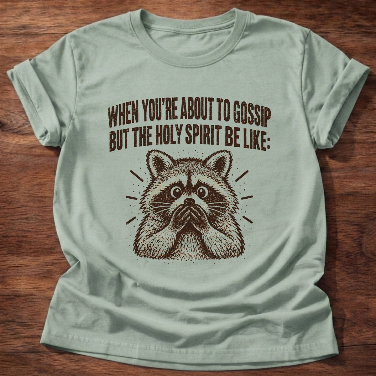Sage t-shirt printed with a startled raccoon graphic and 'When you're about to gossip but the Holy Spirit be like.' Proverbs 16:28 faith based gift