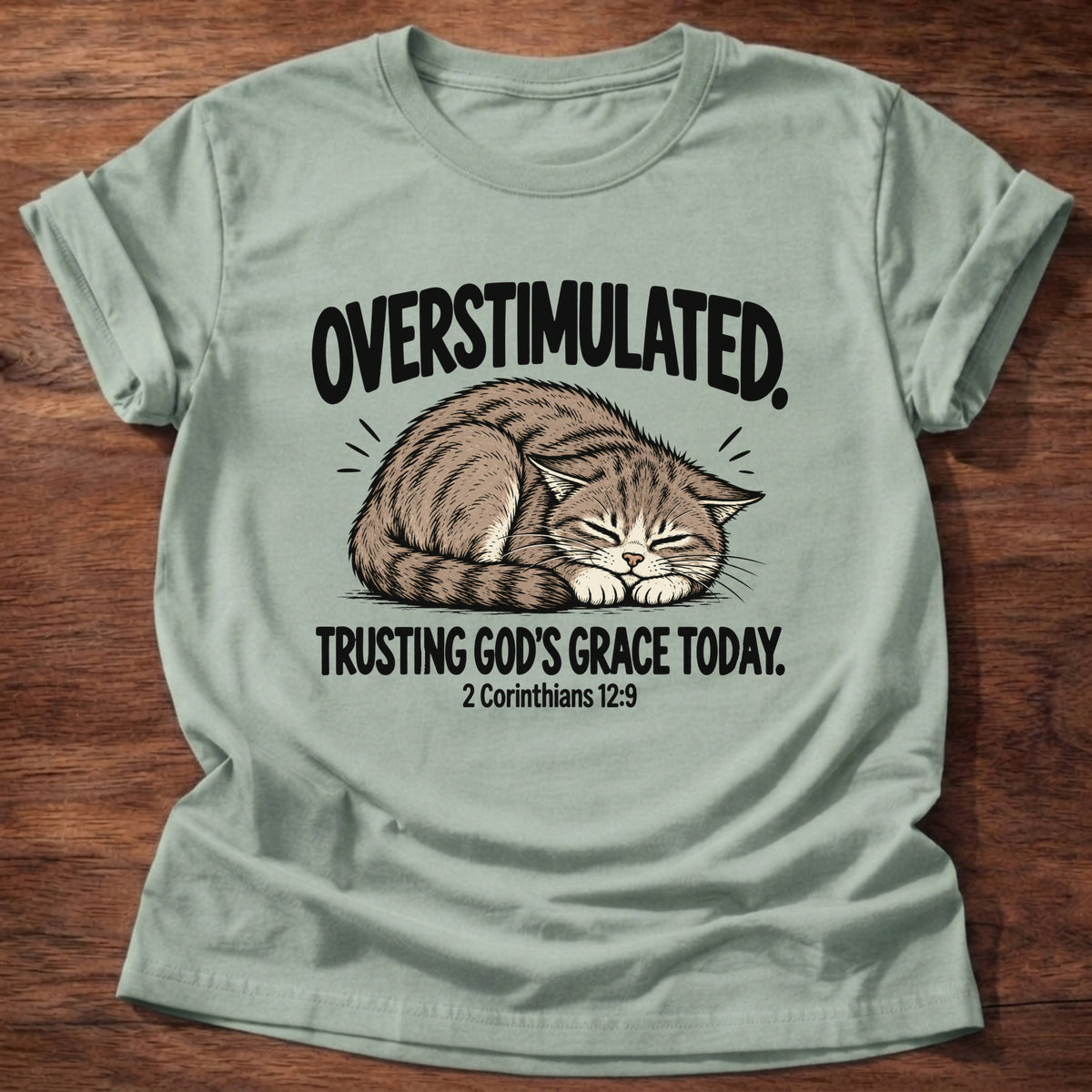 Sage t-shirt printed with a cat curled asleep graphic and 'Overstimulated. Trusting God's grace today.' 2 Corinthians 12:9 faith based gift