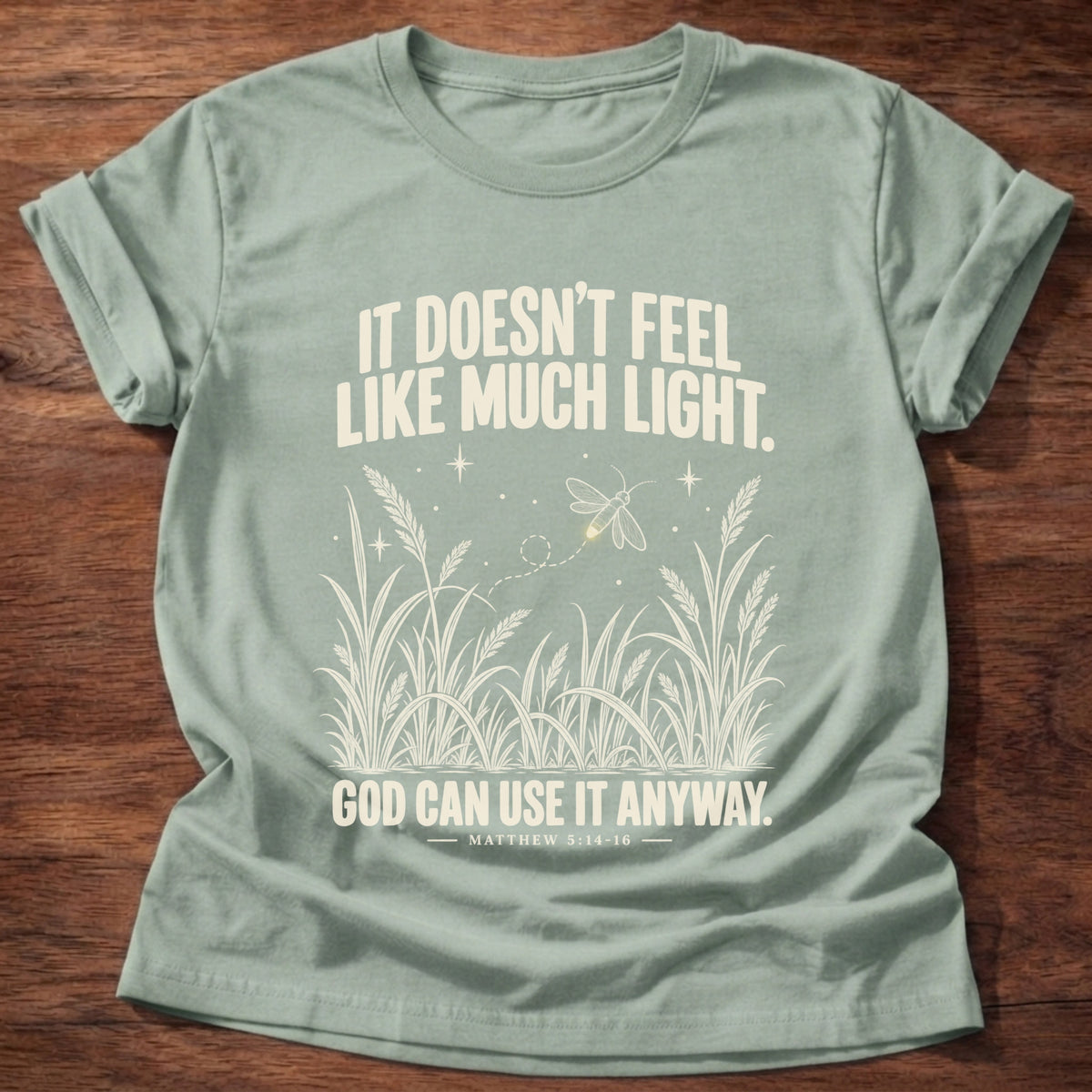 Sage t-shirt printed with a firefly glowing in a dark field of grass graphic and 'It doesn't feel like much light. God can use it anyway.' Matthew 5:14-16 faith based gift