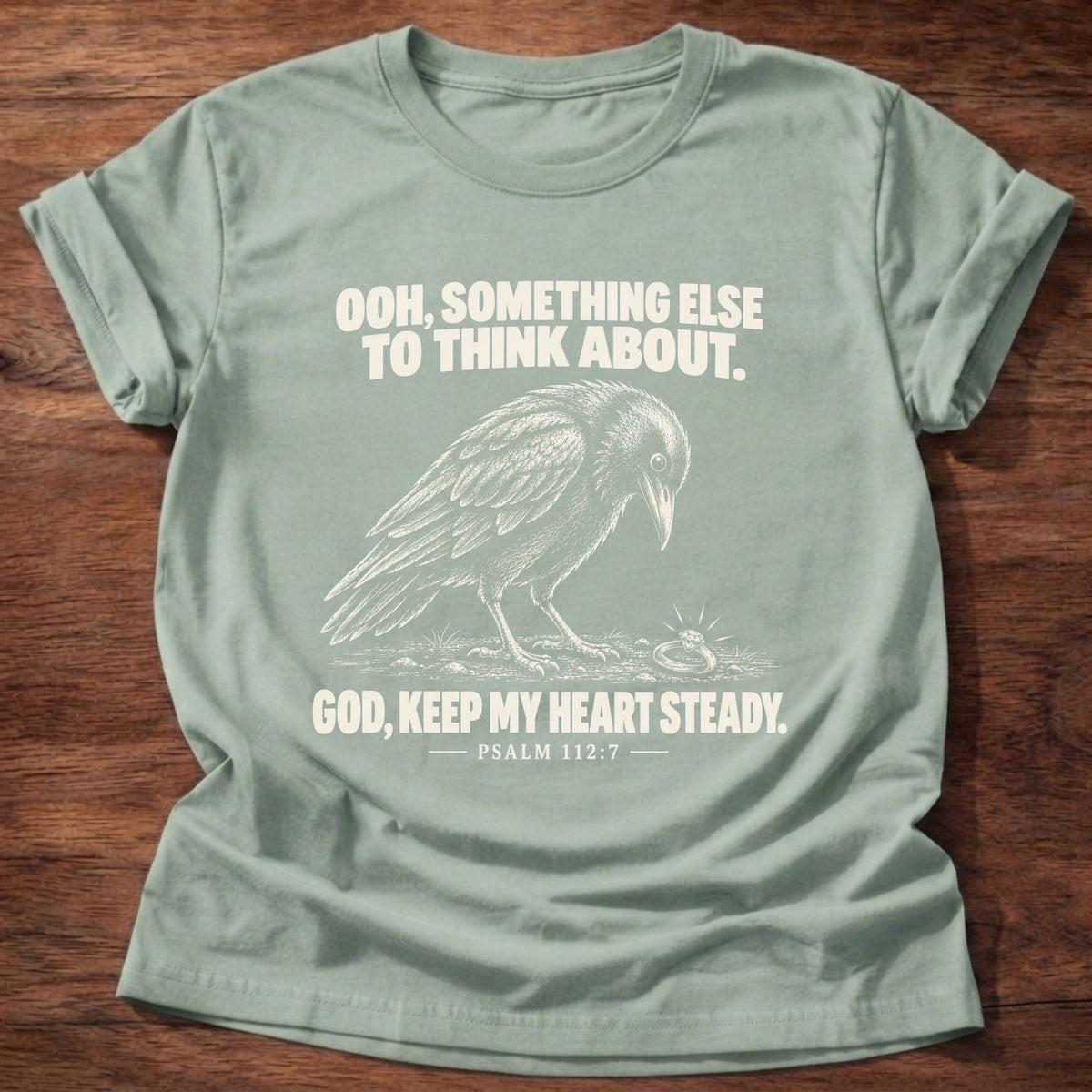 Sage t-shirt printed with a crow investigating something shiny on the ground graphic and 'Ooh, something else to think about. God, keep my heart steady.' Psalm 112:7 faith based gift