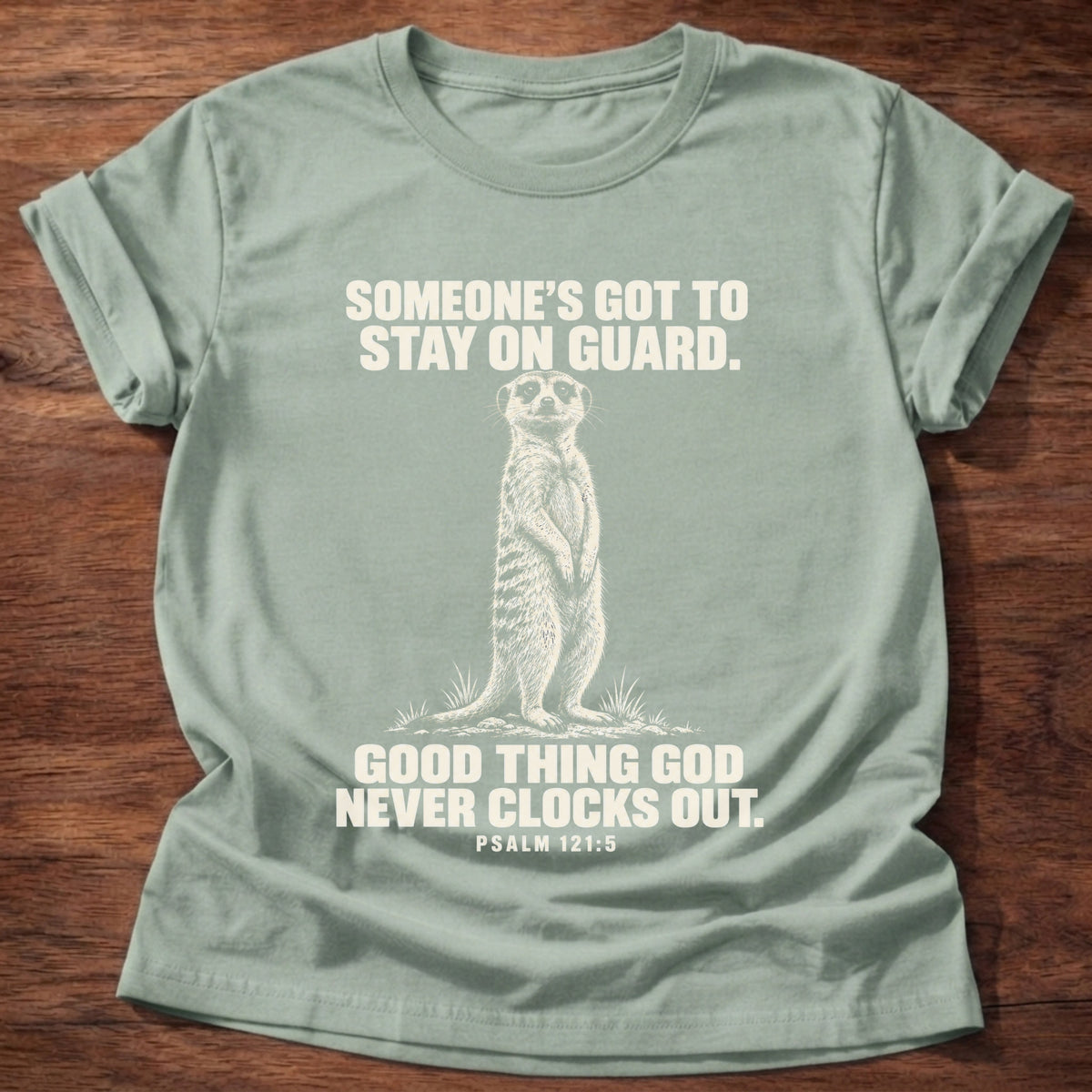Sage t-shirt printed with a meerkat standing guard on lookout graphic and 'Someone's got to stay on guard. Good thing God never clocks out.' Psalm 121:5 faith based gift