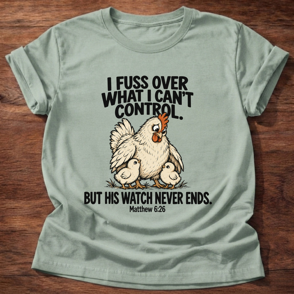 Sage t-shirt printed with a fussing hen graphic and 'I fuss over what I can't control. But His watch never ends.' Matthew 6:26 faith based gift