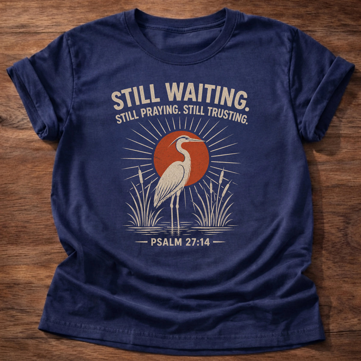 Navy t-shirt with a heron illustration at sunrise reading 'Still waiting. Still praying. Still trusting.' Psalm 27:14 shirt