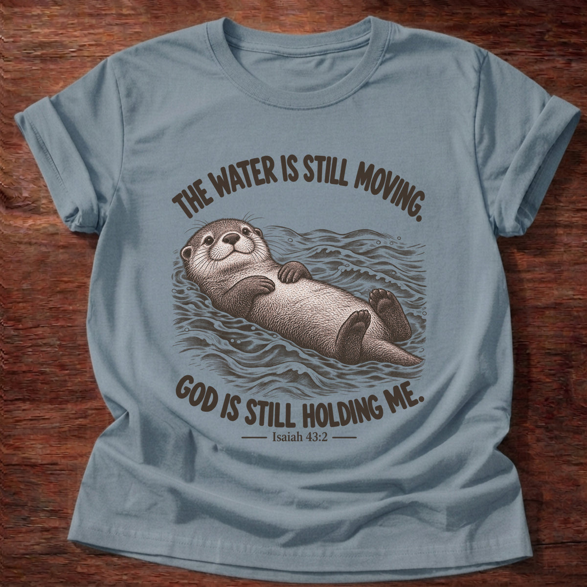 Stone Blue t-shirt printed with an otter floating on its back in moving water graphic and “The water is still moving. God is still holding me.” Isaiah 43:2 faith based gift
