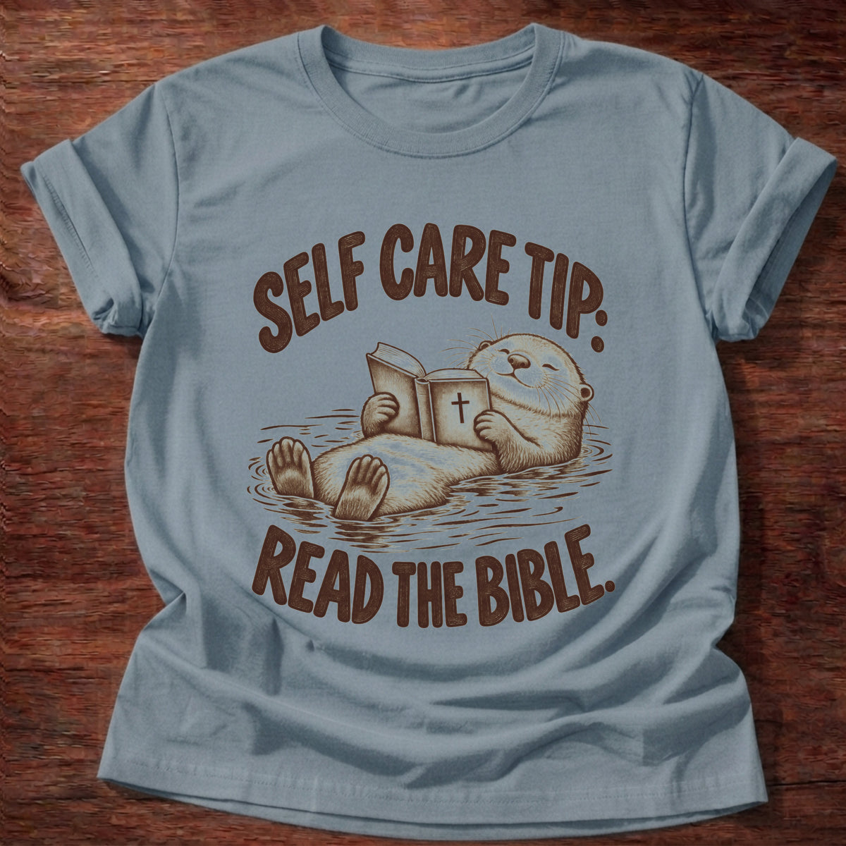 Stone Blue t-shirt printed with a sea otter floating on its back reading a Bible graphic and “Self care tip: Read the Bible.” faith based gift