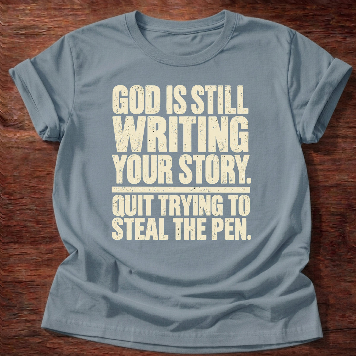 Stone Blue t-shirt printed with bold stacked typography graphic and “God is still writing your story. Quit trying to steal the pen.” Philippians 1:6 Christian shirt