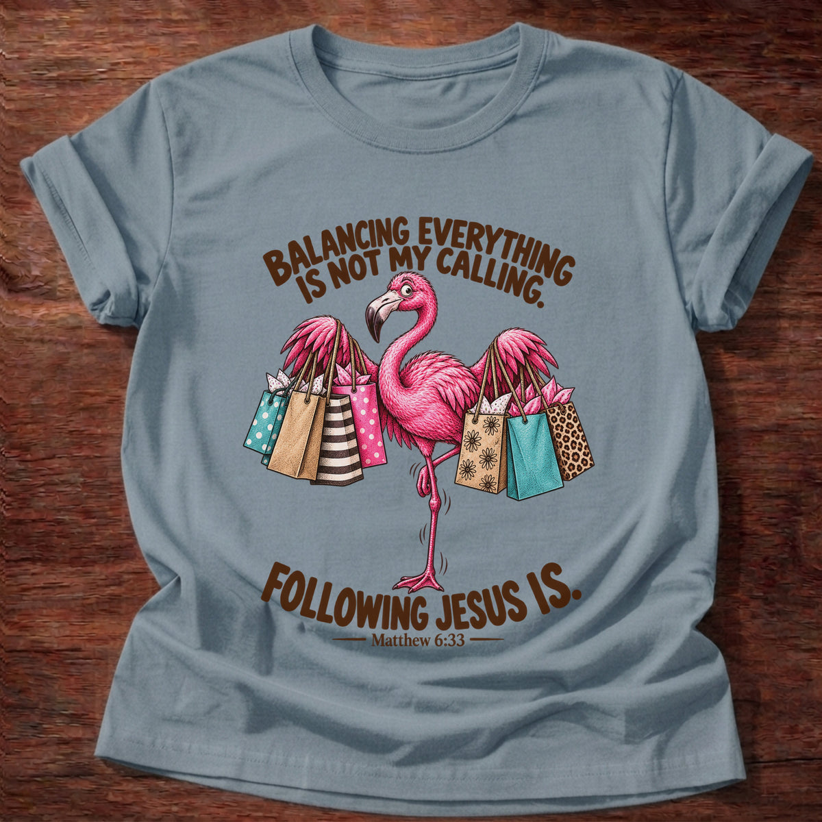 Stone Blue t-shirt printed with a pink flamingo balancing on one leg loaded with colorful shopping bags graphic and “Balancing everything is not my calling. Following Jesus is.” Matthew 6:33 faith based gift
