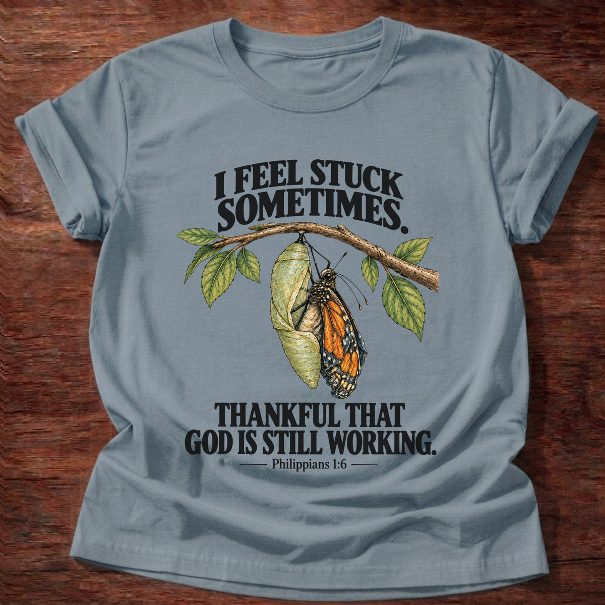 Stone Blue t-shirt printed with a monarch butterfly emerging from its chrysalis on a leafy branch graphic and “I feel stuck sometimes. Thankful that God is still working.” Philippians 1:6 faith based gift
