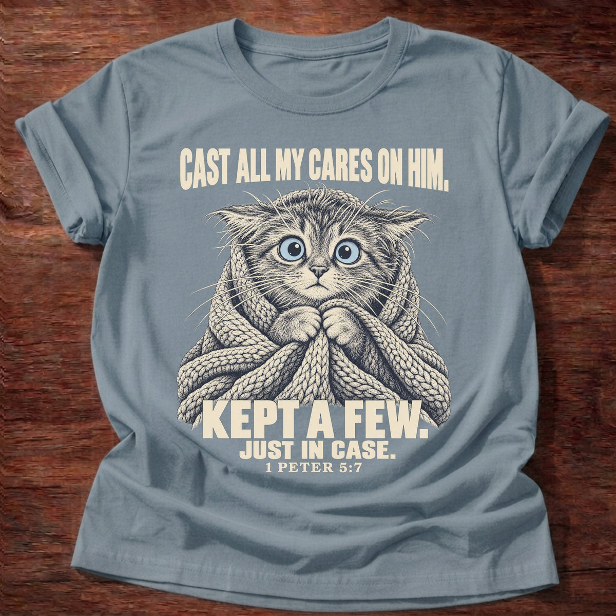 Kept A Few T-Shirt in Stone Blue featuring a wide-eyed cat wrapped in a knit blanket and 1 Peter 5:7 anxiety humor design