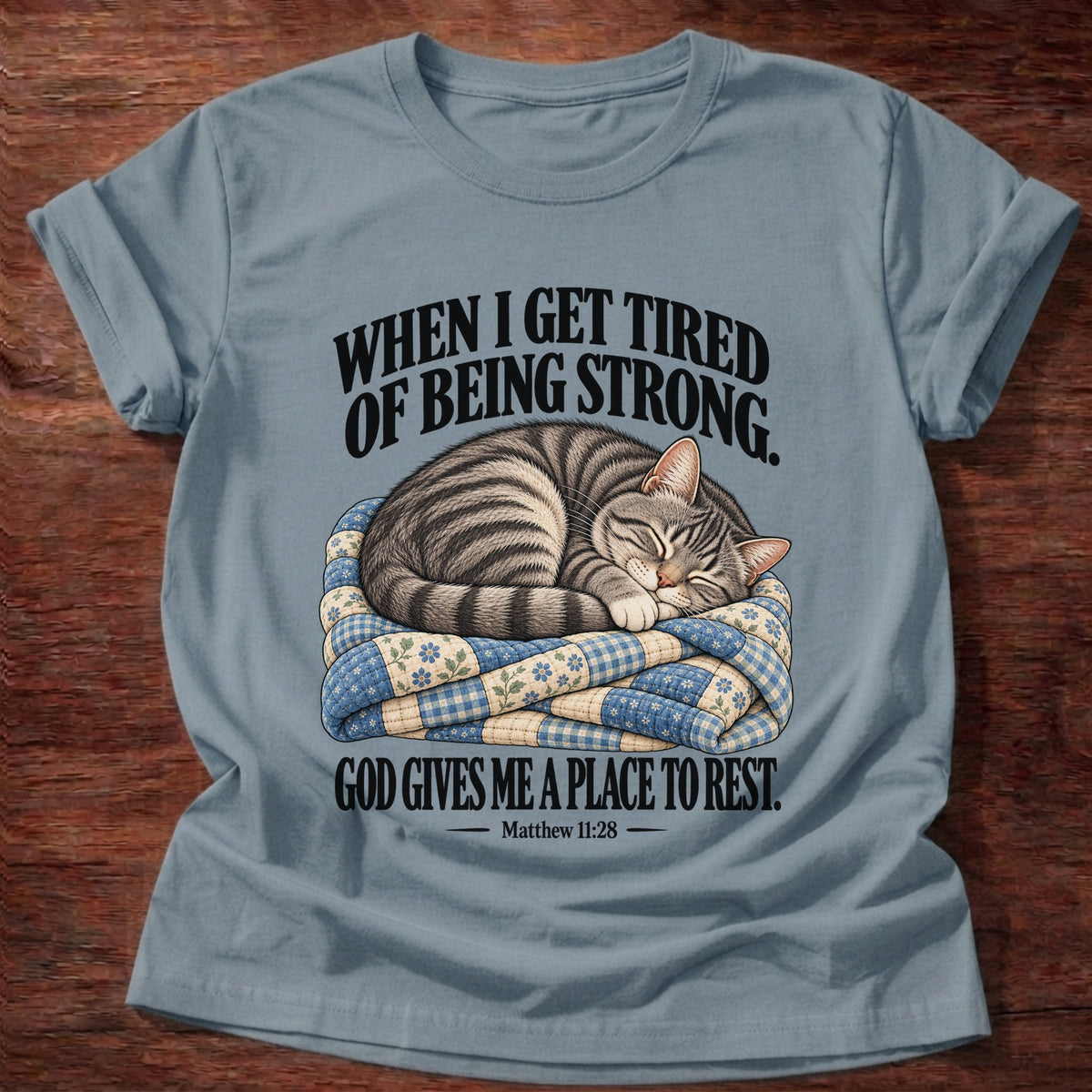 Stone Blue t-shirt printed with a tabby cat curled up asleep on a folded patchwork quilt graphic and “When I get tired of being strong, God gives me a place to rest.” Matthew 11:28 faith based gift