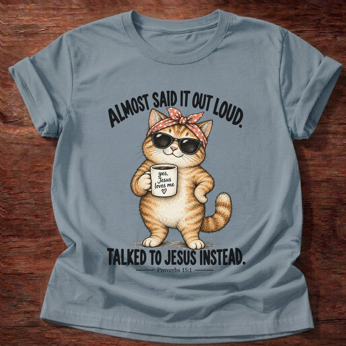 Stone Blue t-shirt printed with an orange tabby cat in sunglasses and a polka-dot bandana holding a 'yes, Jesus loves me' mug graphic and “Almost said it out loud. Talked to Jesus instead.” Proverbs 15:1 faith based gift