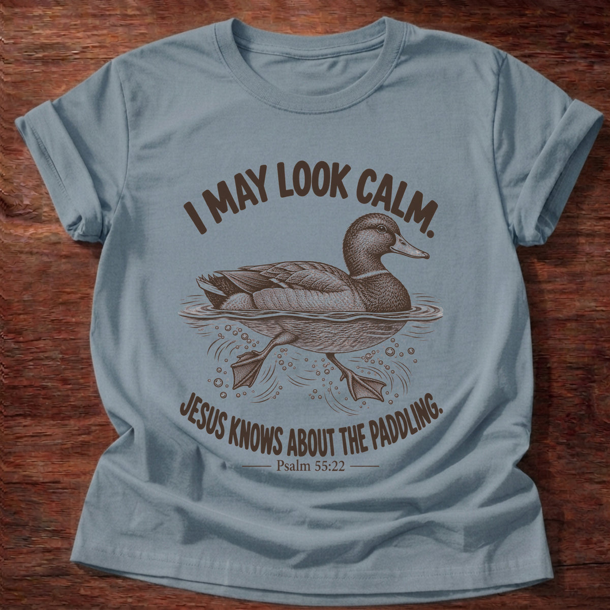 Stone Blue t-shirt printed with a mallard duck gliding on the water with its feet paddling underneath graphic and “I may look calm. Jesus knows about the paddling.” Psalm 55:22 faith based gift