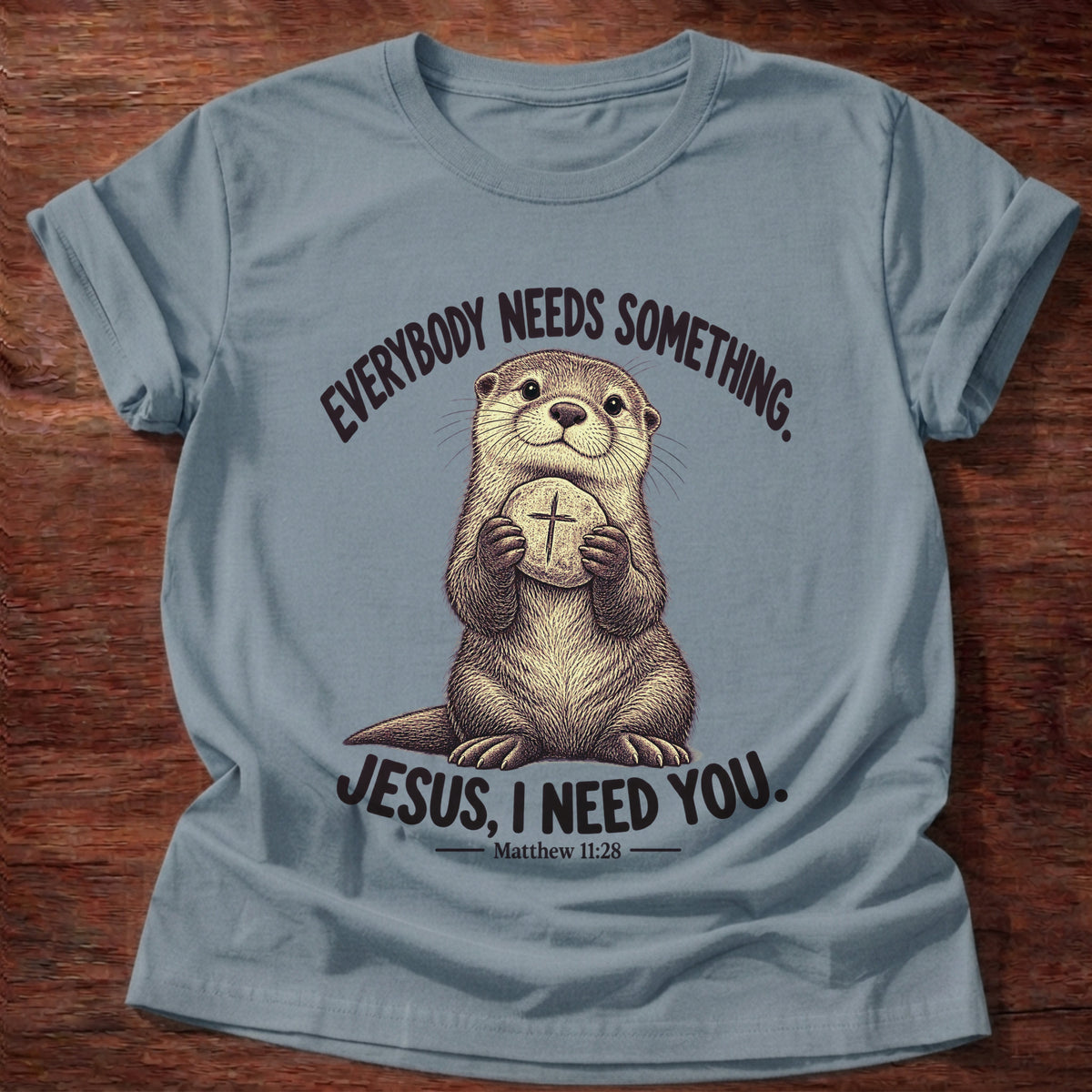 Stone Blue t-shirt printed with an otter hugging a round stone etched with a cross graphic and “Everybody needs something. Jesus, I need you.” Matthew 11:28 faith based gift
