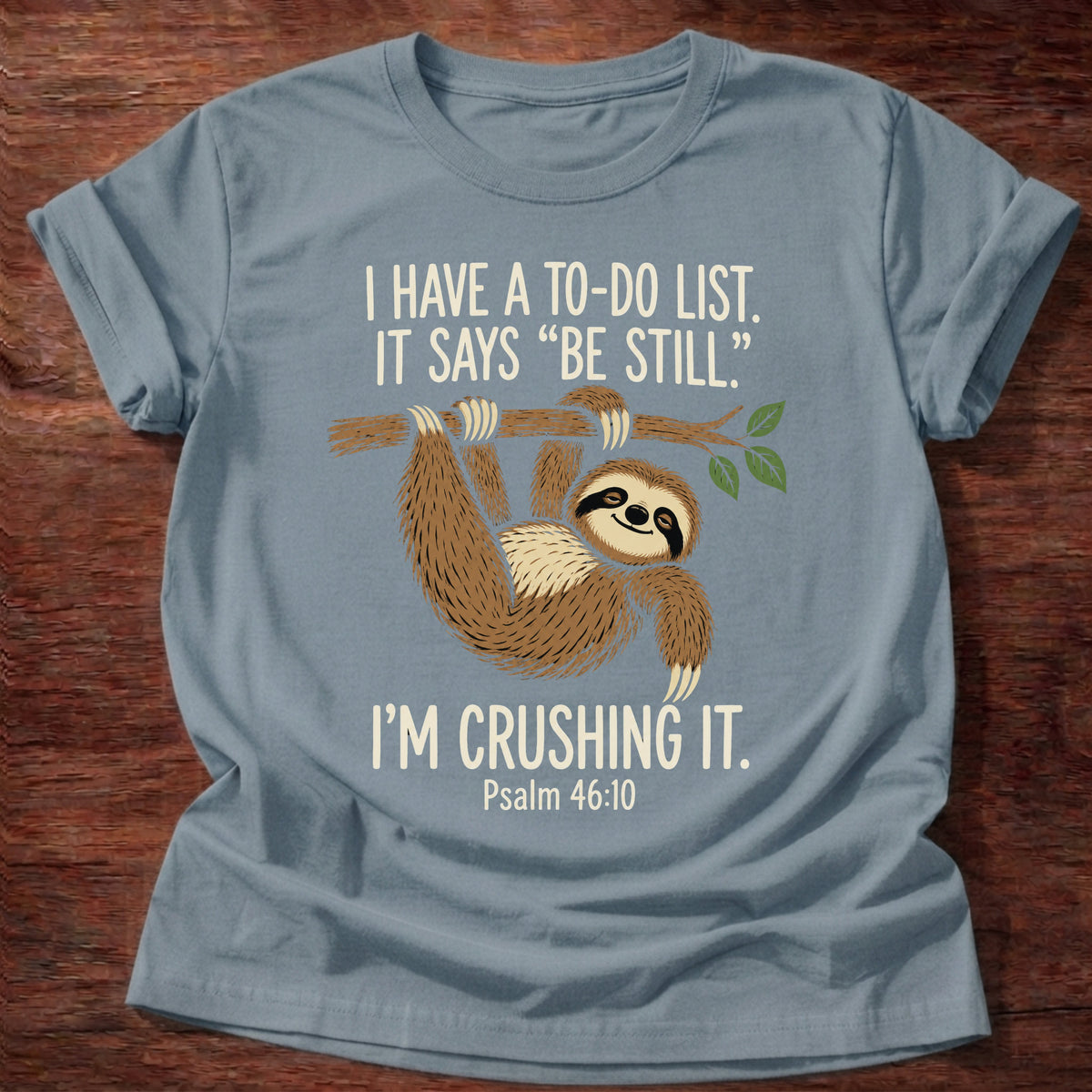Stone Blue t-shirt printed with a happy sloth hanging from a branch graphic and “I have a to-do list. It says 'Be still.' I'm crushing it.” Psalm 46:10 Christian shirt