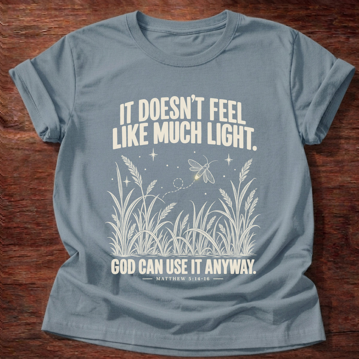 Stone Blue t-shirt printed with a firefly glowing in a dark field of grass graphic and 'It doesn't feel like much light. God can use it anyway.' Matthew 5:14-16 faith based gift