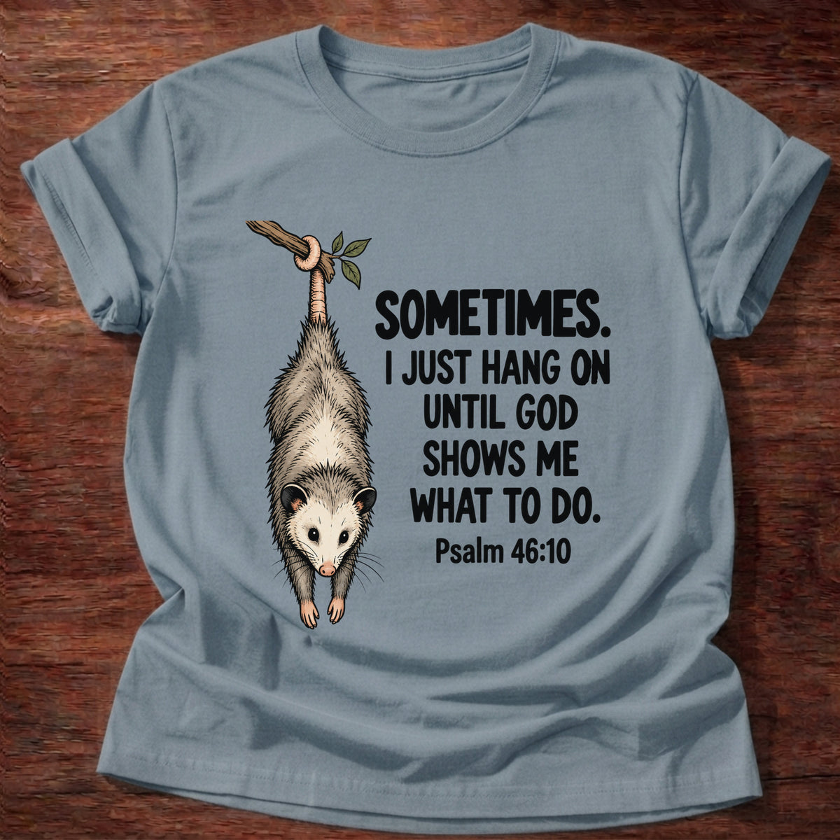 Stone Blue t-shirt printed with a possum hanging by its tail graphic and 'Sometimes. I just hang on until God shows me what to do.' Psalm 46:10 faith based gift