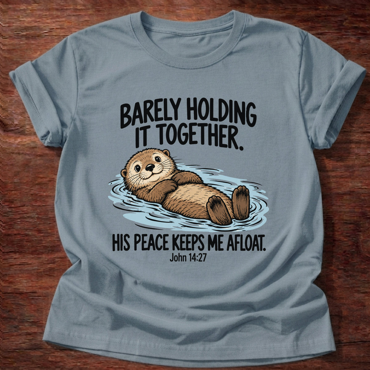 Stone Blue t-shirt printed with an otter floating on its back graphic and 'Barely holding it together. His peace keeps me afloat.' John 14:27 faith based gift