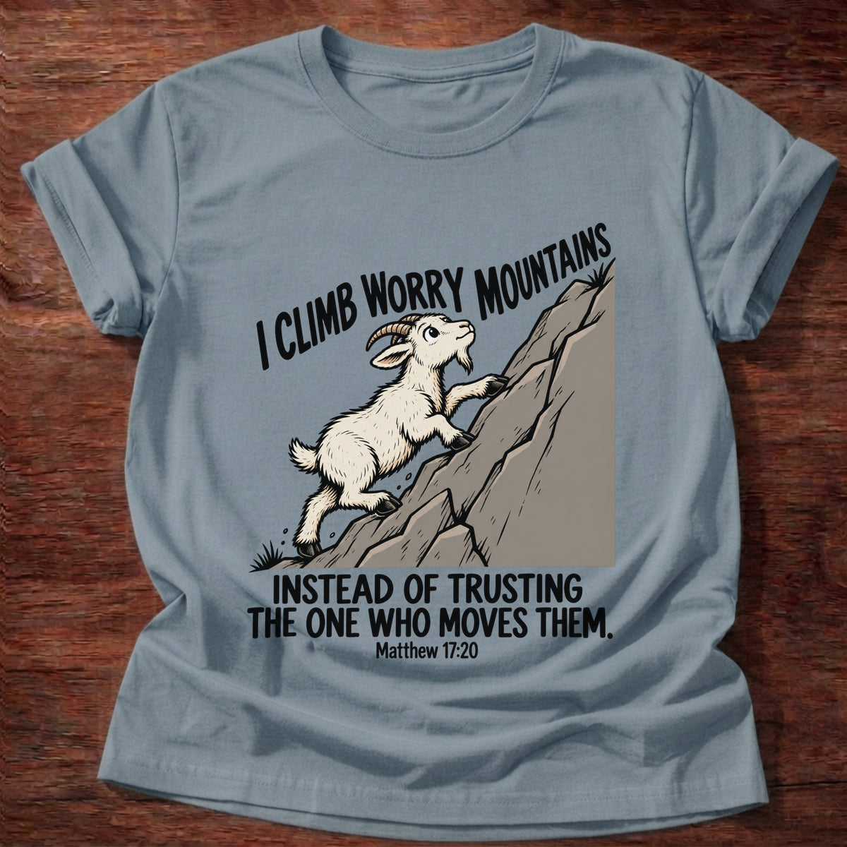 Stone Blue t-shirt printed with a goat climbing a mountain graphic and 'I climb worry mountains instead of trusting the One who moves them.' Matthew 17:20 faith based gift