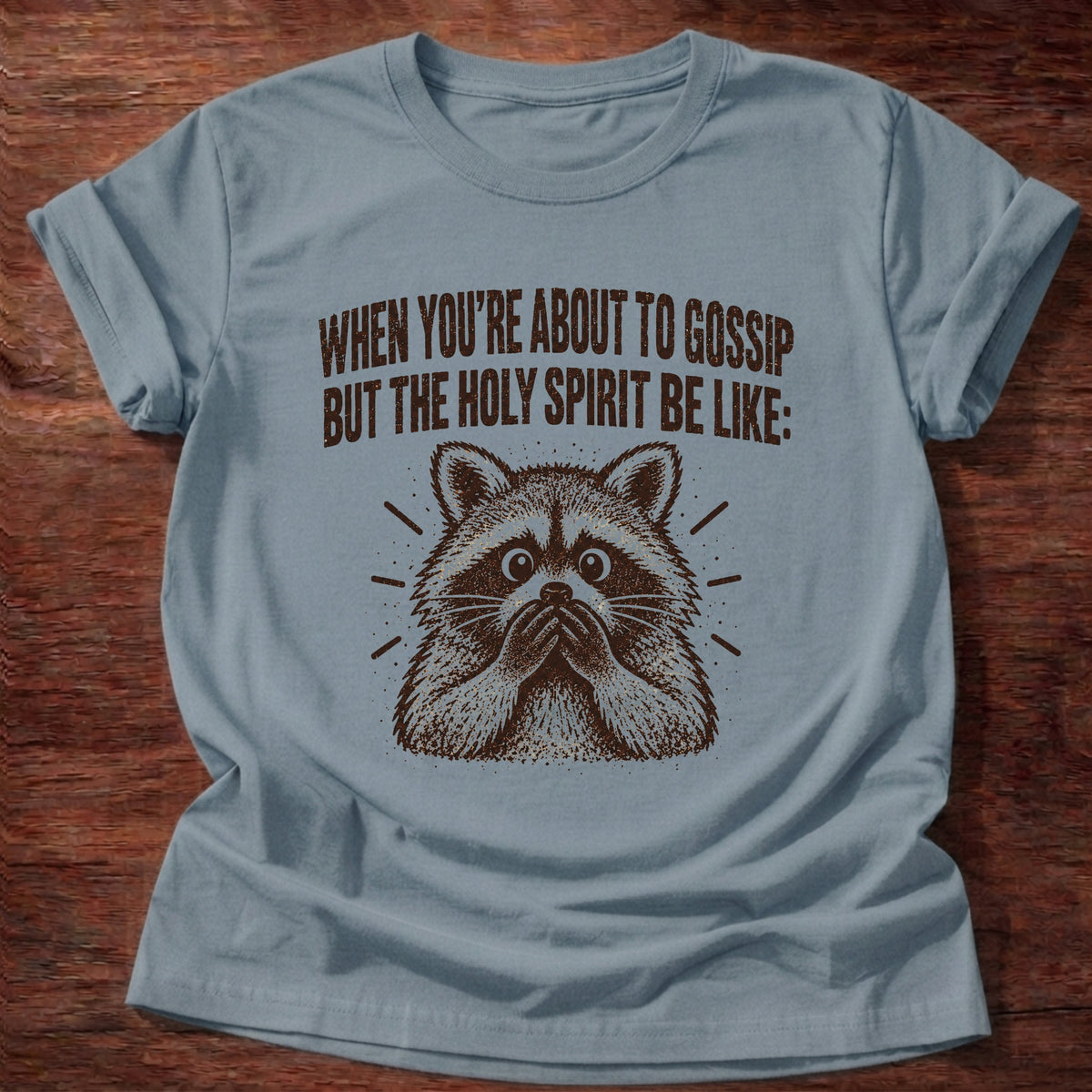 Stone Blue t-shirt printed with a startled raccoon graphic and 'When you're about to gossip but the Holy Spirit be like.' Proverbs 16:28 faith based gift