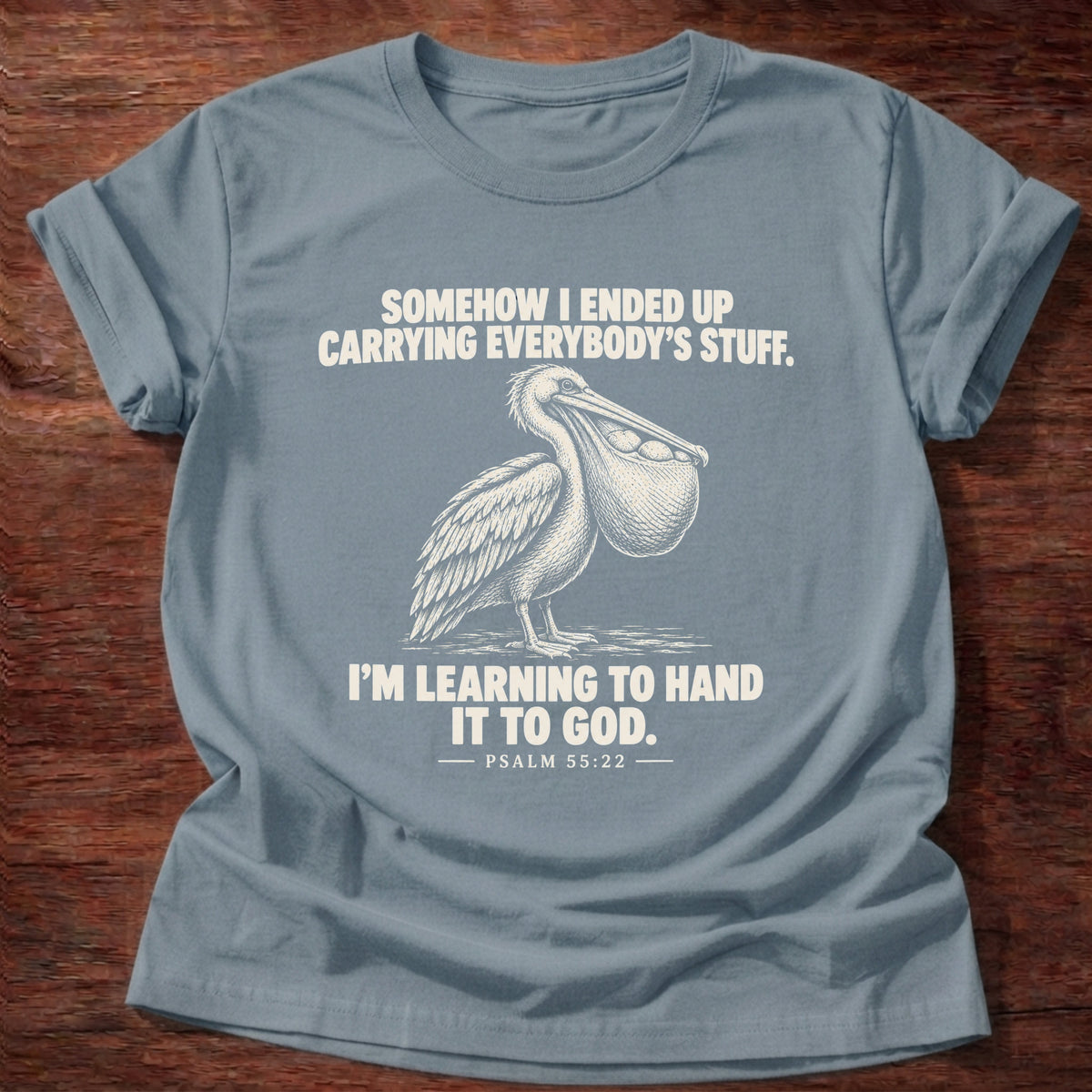Stone Blue t-shirt printed with a pelican with a pouch full of fish graphic and 'Somehow I ended up carrying everybody's stuff. I'm learning to hand it to God.' Psalm 55:22 faith based gift