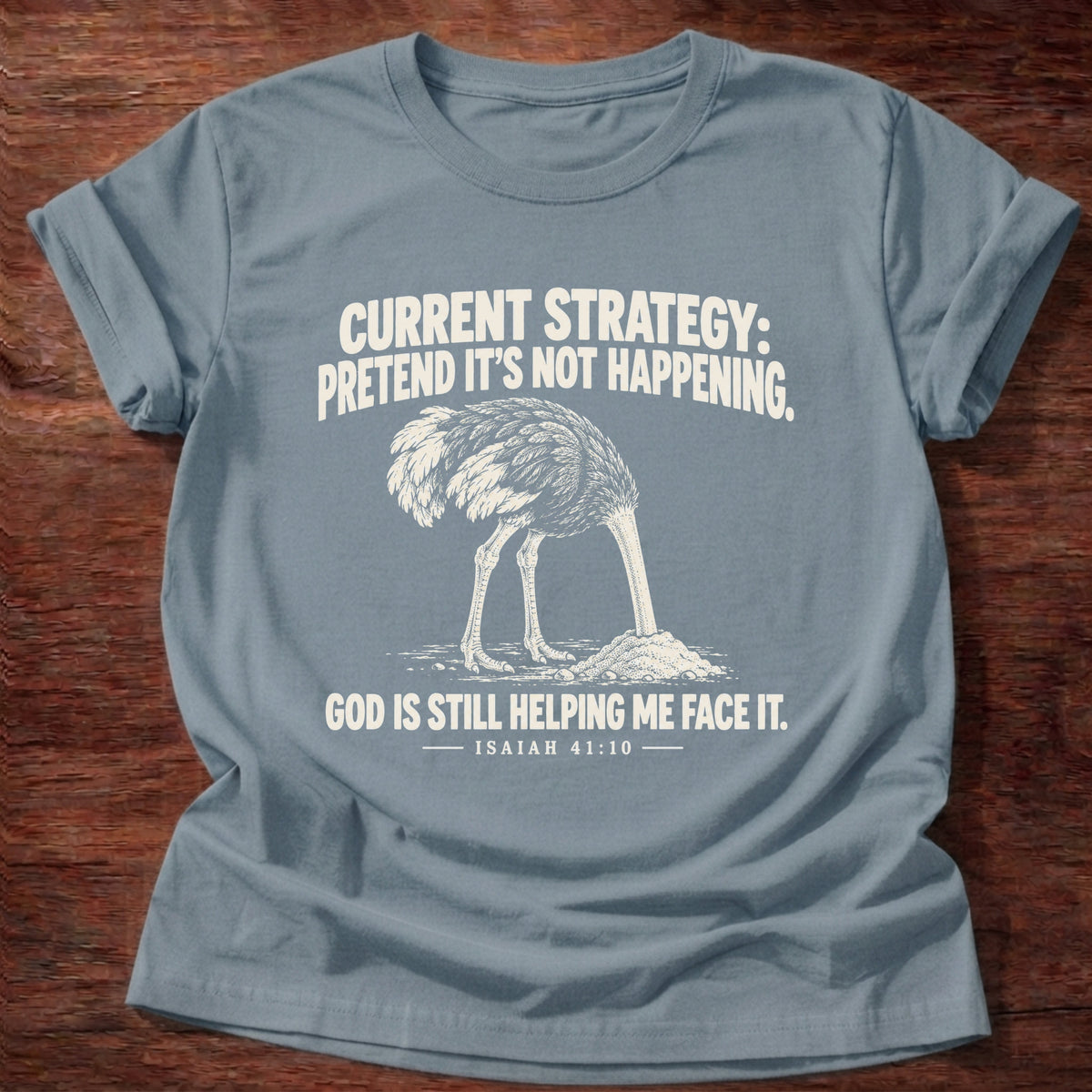 Stone Blue t-shirt printed with an ostrich with its head buried in the sand graphic and 'Current strategy: pretend it's not happening. God is still helping me face it.' Isaiah 41:10 faith based gift