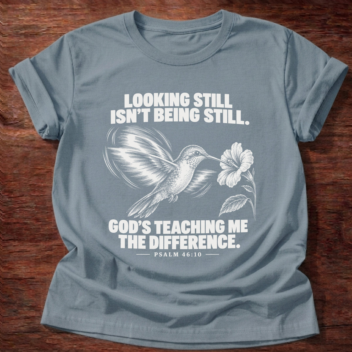 Stone Blue t-shirt printed with a hummingbird hovering beside a flower graphic and 'Looking still isn't being still. God's teaching me the difference.' Psalm 46:10 faith based gift