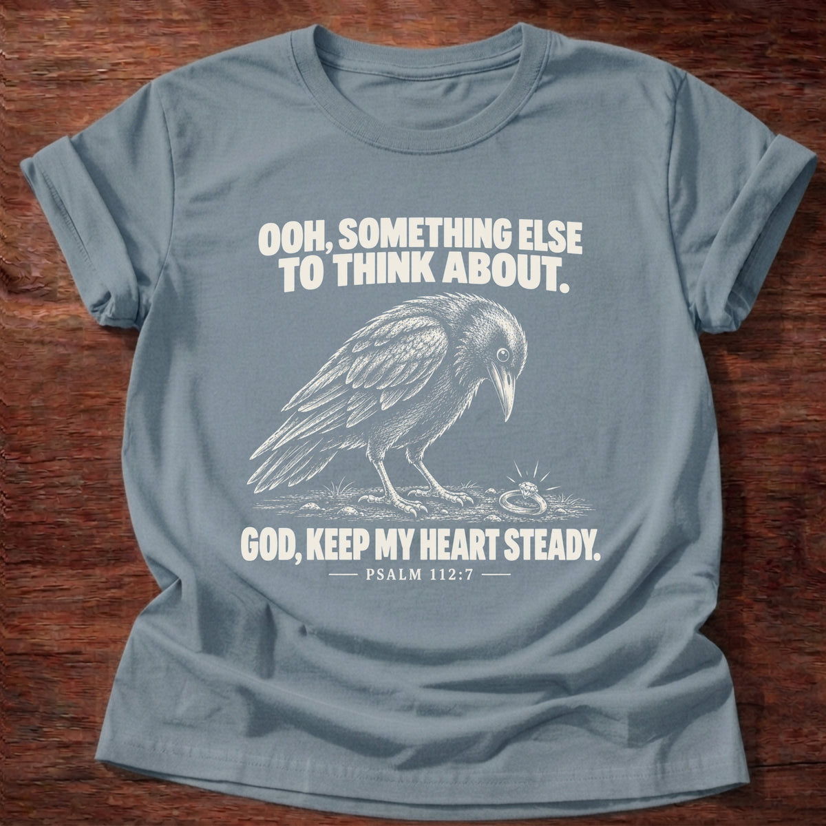 Stone Blue t-shirt printed with a crow investigating something shiny on the ground graphic and 'Ooh, something else to think about. God, keep my heart steady.' Psalm 112:7 faith based gift