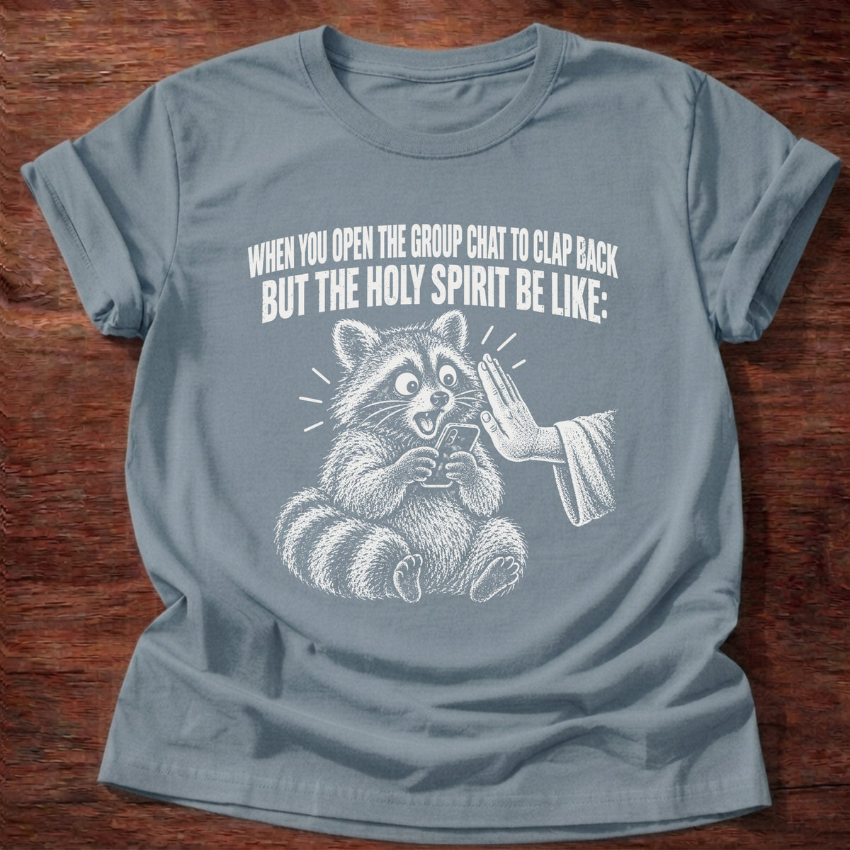 Stone Blue t-shirt printed with a raccoon covering its own mouth graphic and 'When you open the group chat to clap back but the Holy Spirit be like.' Proverbs 21:23 faith based gift