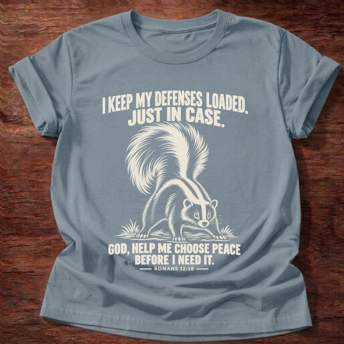 Stone Blue t-shirt printed with a skunk with its tail raised, ready to spray graphic and 'I keep my defenses loaded. Just in case. God, help me choose peace before I need it.' Romans 12:18 faith based gift