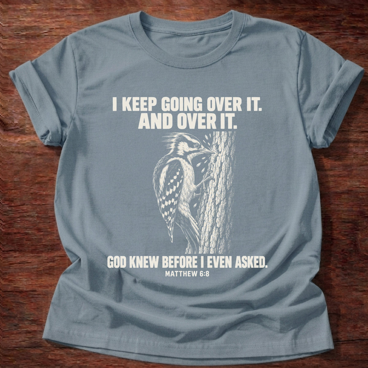 Stone Blue t-shirt printed with a woodpecker drumming the same spot on a tree graphic and 'I keep going over it. And over it. God knew before I even asked.' Matthew 6:8 faith based gift