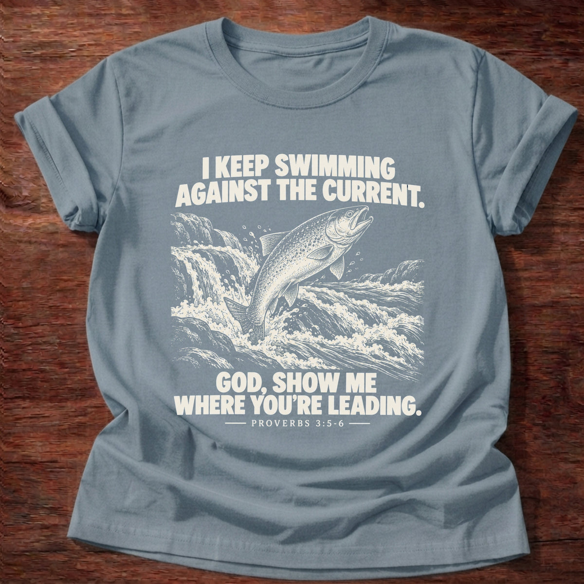 Stone Blue t-shirt printed with a fish leaping against a rushing current graphic and 'I keep swimming against the current. God, show me where you're leading.' Proverbs 3:5-6 faith based gift