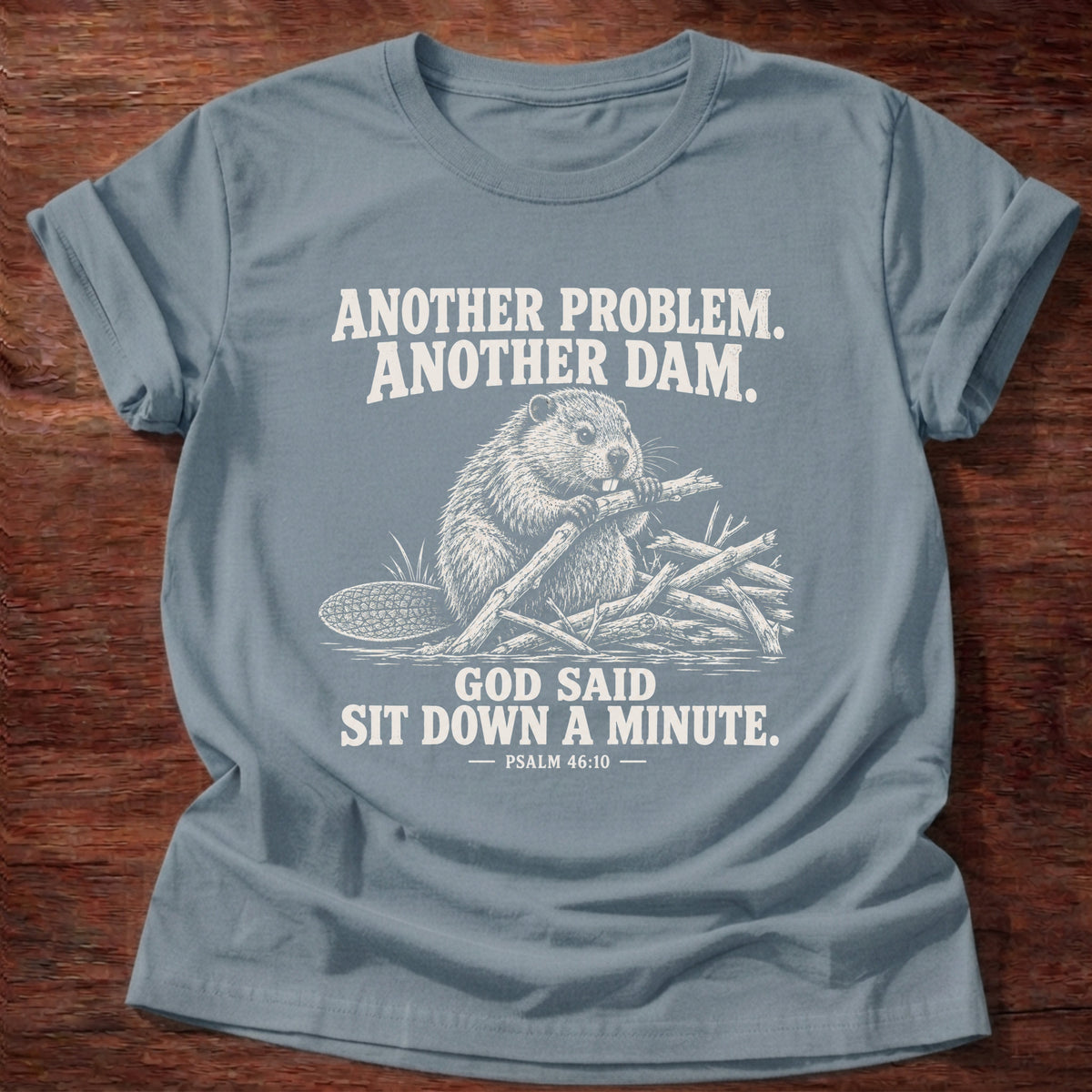 Stone Blue t-shirt printed with a beaver mid-build with another branch graphic and 'Another problem. Another dam. God said sit down a minute.' Psalm 46:10 faith based gift