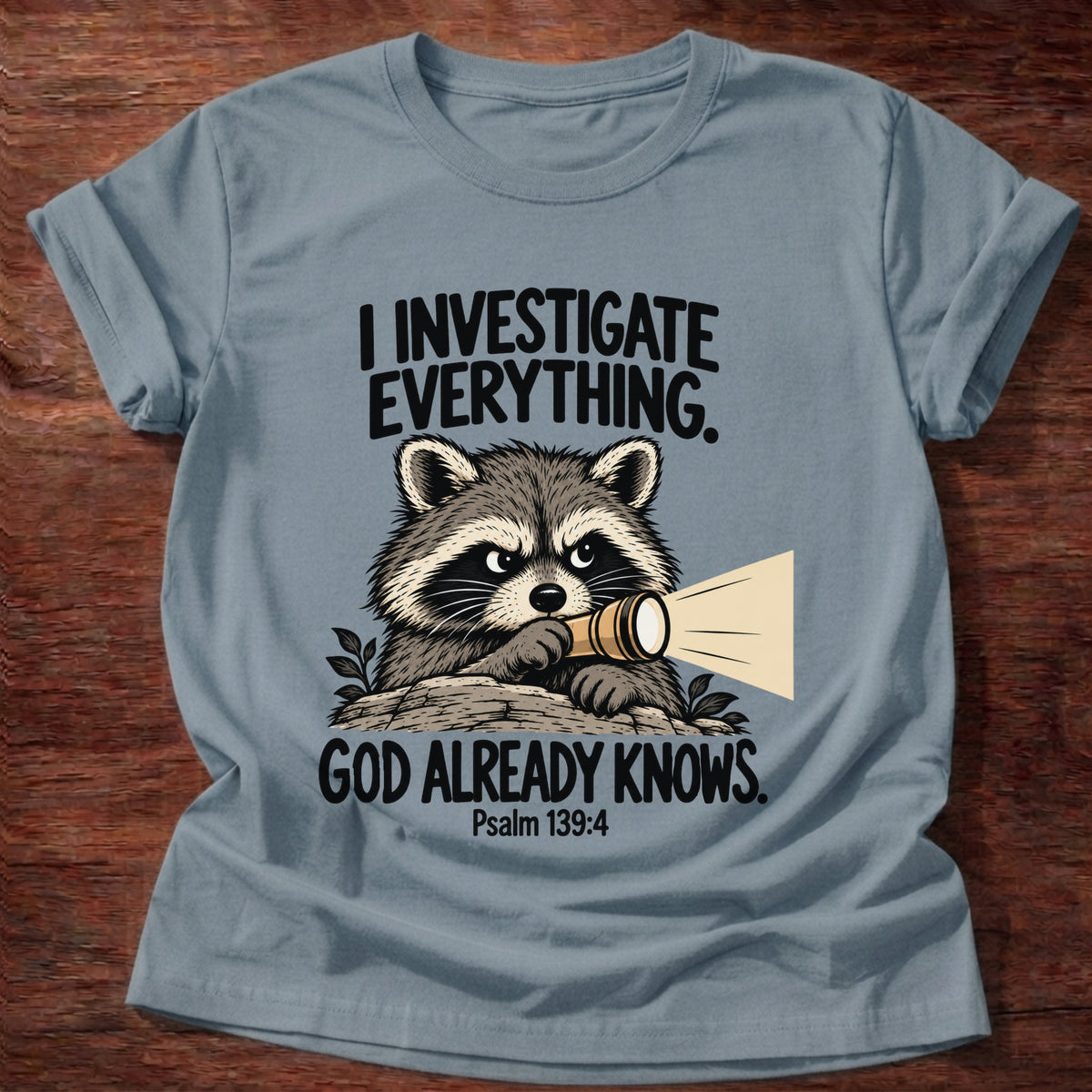 Stone Blue t-shirt printed with a raccoon holding a magnifying glass graphic and 'I investigate everything. God already knows.' Psalm 139:4 faith based gift